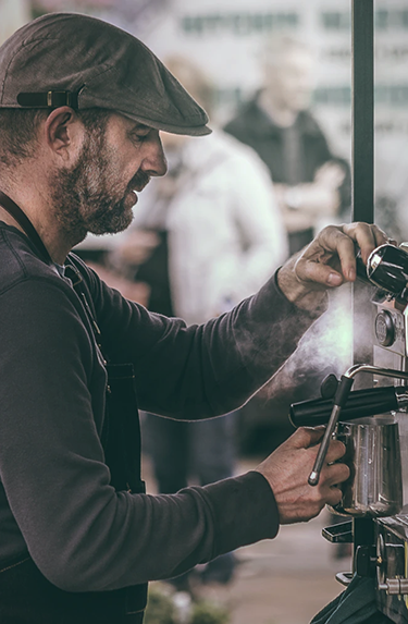 An older, male barista steaming milk in a cafe wearing a cabbie hat and a long sleeved henley.