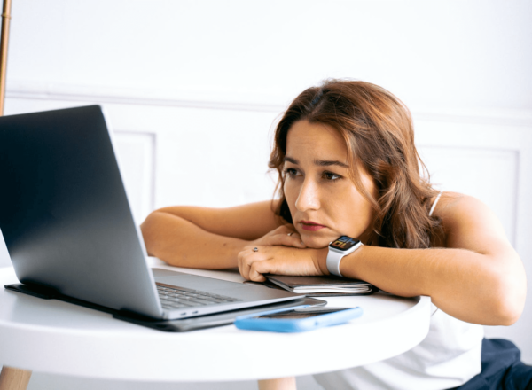 Image of woman sitting at a desk with her arms on the table looking slightly anxious at her laptop screen.