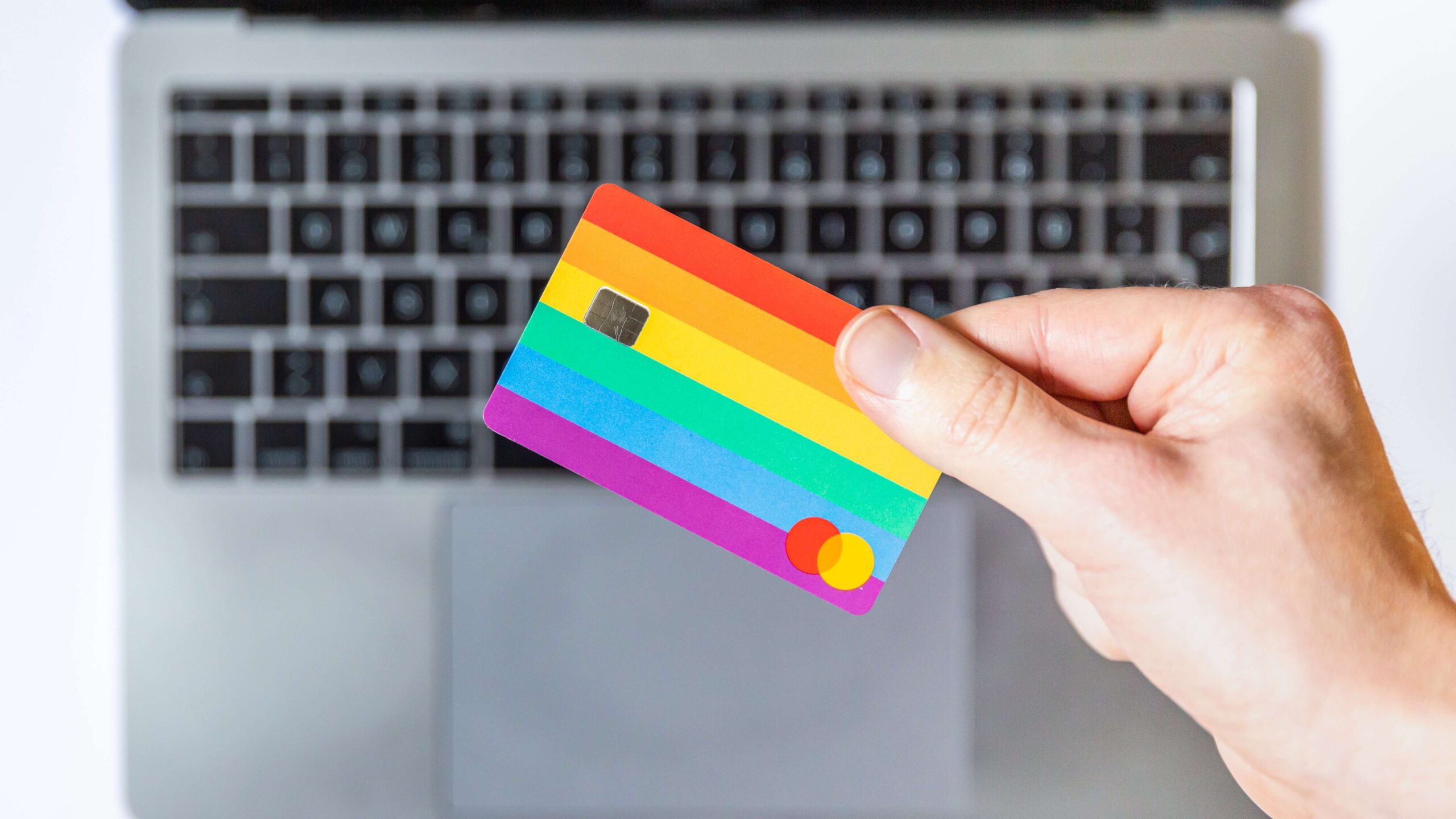 A person's hand holding an LGBT themed credit card in front of a laptop.